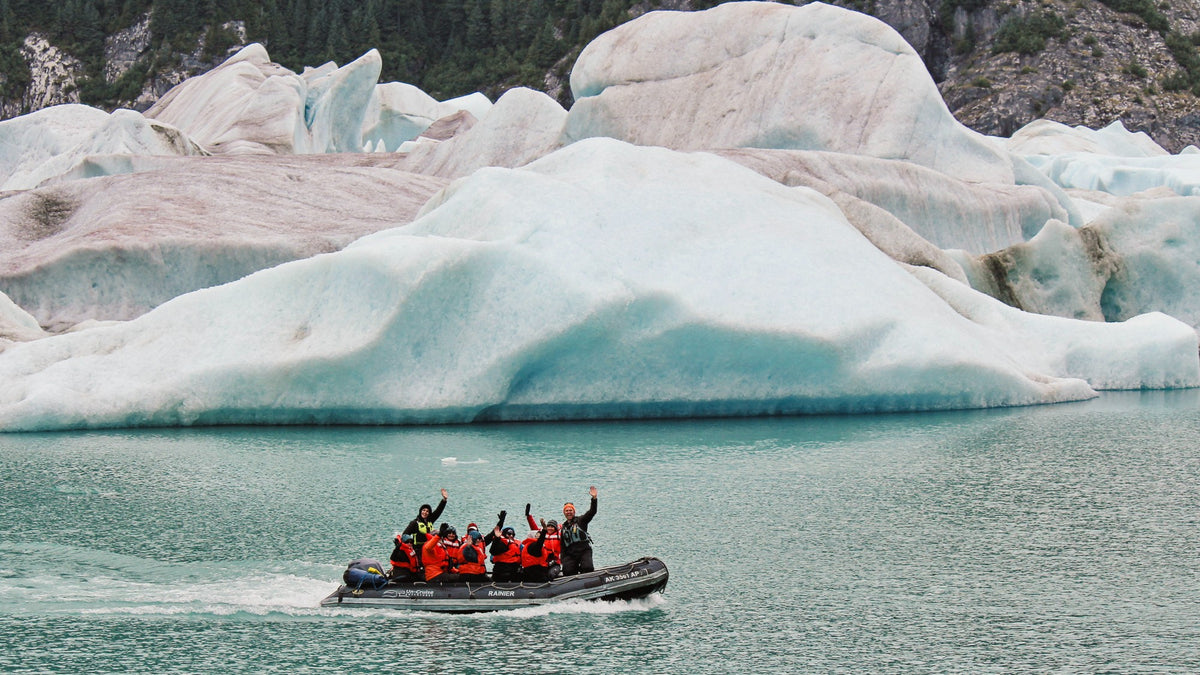 Skiff full of guests waiving at the camera as it motors past a large iceberg