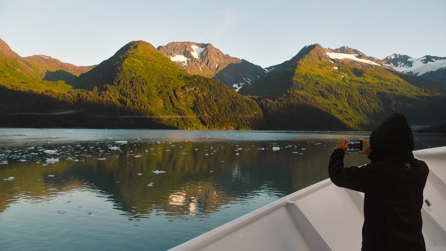 A person in a black hoodie stands on the edge of a boat, holding a smartphone to take a photo of a scenic mountain with green slopes, snow patches, and calm water under a clear sky.