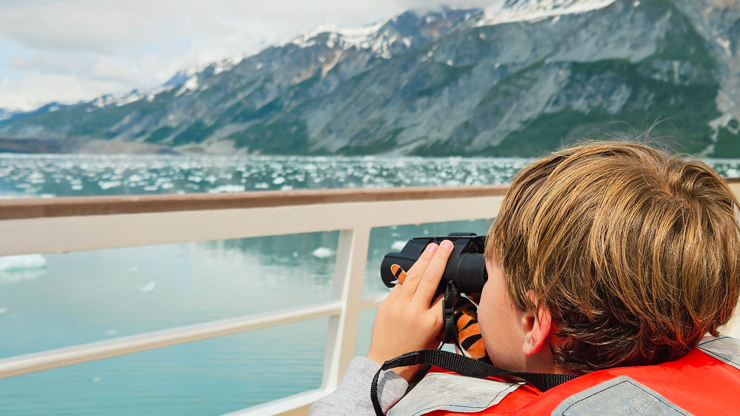 A child in a red life jacket looks through binoculars from a boat deck, with a scenic mountain and glacial fjord in the background.