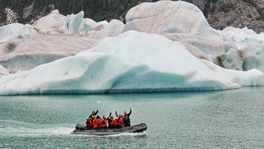 Skiff full of guests waiving at the camera as it motors past a large iceberg-UnCruise Adventures 