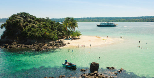 Scenic tropical beach with turquoise water, a small sandy cove, a rocky green island, palm trees, swimmers, a small boat, and a larger ferry in the distance under a clear blue sky.-UnCruise Adventures 