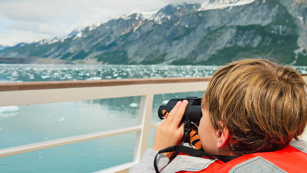 A child in a red life jacket looks through binoculars from a boat deck, with a scenic mountain and glacial fjord in the background.