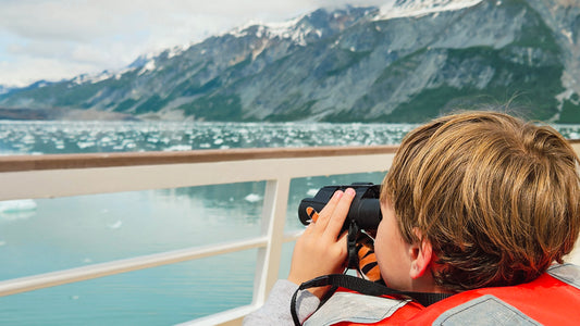 A child in a red life jacket looks through binoculars from a boat deck, with a scenic mountain and glacial fjord in the background.-UnCruise Adventures 
