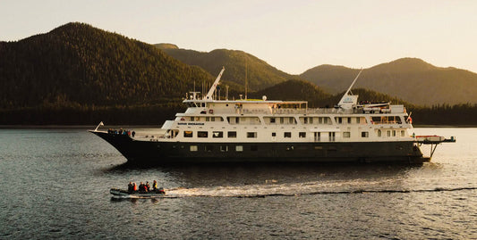 Expedition ship sails past mountains at sunset as skiff drives beside it-UnCruise Adventures 