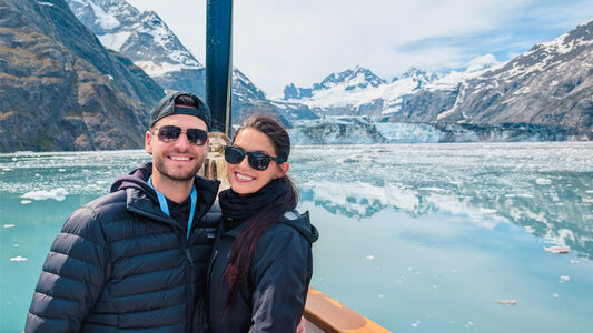 A smiling couple posing on a boat in front of a glacier fjord with snow-capped mountains in the background. -UnCruise Adventures 